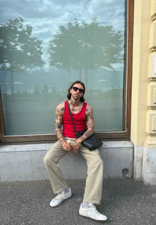 Hombre tatuado con camiseta de tirantes roja, pantalones beige y gafas de sol, sentado en una repisa, sosteniendo un cucurucho de helado, con árboles reflejados en la gran ventana detrás de él.