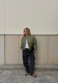 Green bomber jacket, grey cardigan, white t-shirt, black cargo trousers, and leopard print flats. Sunglasses and a small black handbag included.