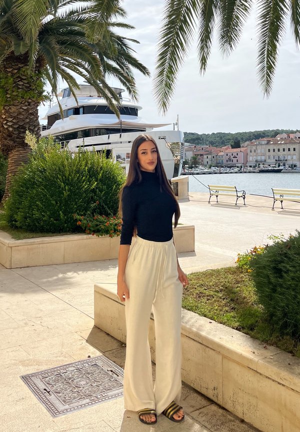 Jeune femme en haut noir et pantalon blanc, debout près de palmiers au bord d'une marina avec un grand yacht et des bancs en arrière-plan.
