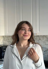 Camicia di raso bianco con piping nero, collane d'oro a strati con charm geometrici, luce morbida e sfondo in marmo sottile.