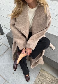 Beige, textured oversized coat with a shawl collar, layered over a cream sweater and black trousers, teamed with dark brown flats.