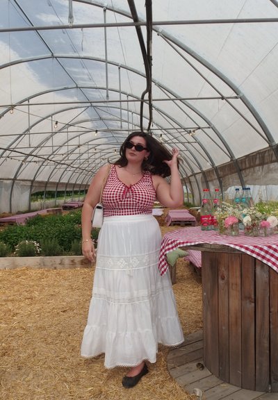 Top corto a cuadros rojos y blancos con un lazo, combinado con una falda larga blanca con detalles de encaje. Zapatos planos negros. Entorno de invernadero.