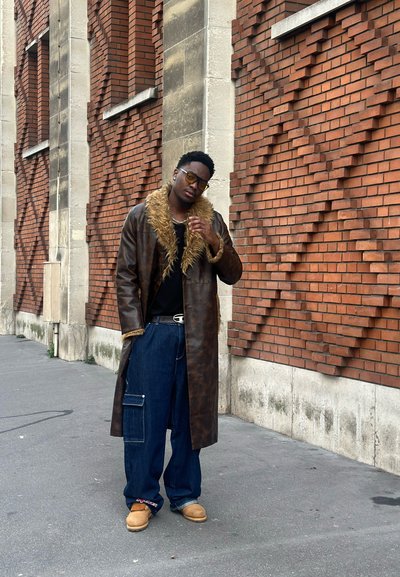 Abrigo de cuero marrón con cuello de piel sintética, camisa negra y jeans azules holgados con bolsillos. De pie en una calle contra una pared de ladrillos.