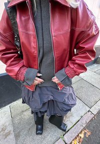 Crvena kožna jakna sa bijelim prošivima, sivi teksturirani džemper, siva plisirana suknja i crne gležnjače, s višeslojnim manžetama i dodacima.