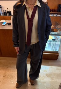 Personne portant un costume gris foncé, un gilet en tricot crème, une chemise rayée, une cravate bordeaux nouée lâchement et des chaussures brunes, debout dans un café près d'une vitrine de pâtisseries.