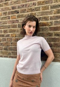 Jonge vrouw met kort bruin haar, gekleed in een lichtroze top met korte mouwen en een suède rok met kralenversieringen, staande tegen een bakstenen muur.