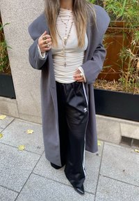 Manteau gris surdimensionné, haut blanc ajusté et pantalons de jogging noirs avec des rayures blanches. Des bijoux métalliques et des chaussures noires pointues complètent le look.