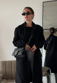 Femme portant un manteau noir, des lunettes de soleil, un pantalon gris et tenant un sac à main noir avec une fermeture dorée, debout à l'intérieur près d'un miroir et d'un canapé.