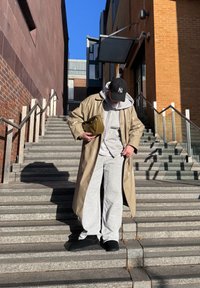 Personne en trench-coat beige, sweat-shirt à capuche gris, pantalon de jogging et casquette noire, debout sur des marches en pierre en plein air, tenant une pochette vert olive.