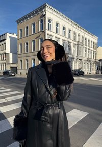 Fekete bőr trench kabát bundával díszített mandzsettákkal, hozzáillő fekete fülvédőkkel és kesztyűkkel; egy v�árosi utcán áll egy történelmi épület közelében.