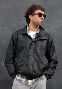 Άνδρας με σγουρά μαλλιά φορώντας μαύρα γυαλιά ηλίου, μαύρο δερμάτινο μπουφάν πάνω από ένα καρό πουκάμισο και ανοιχτό γκρι παντελόνι, που στέκεται μπροστά σε έναν σκουρόχρωμο γκρίζο τοίχο.