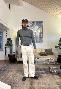 Model szürke, testhez simuló garbót és bő fehér farmert visel, fekete edzőcipőkkel és fekete sapkával. A szobában kanapé és növények találhatók.