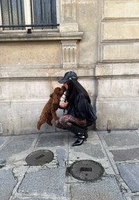 Kvinna i svart klädsel hukar på stadens trottoar och kysser en liten brun hund som står på bakbenen mot en stenvägg.