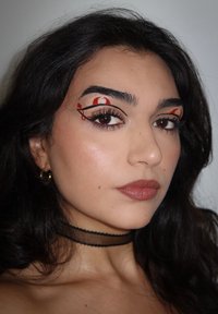 Kobieta z długimi ciemnymi włosami nosi czarną choker oraz złote kolczyki w kształcie kół, a na brązowych oczach ma artystyczne wzory czerwonego i czarnego eyelineru.