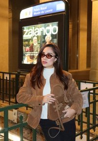 Brun mockajacka med en ljus polotröja, kombinerad med mörka byxor. Accessoarerna inkluderar en liten handväska och runda solglasögon.