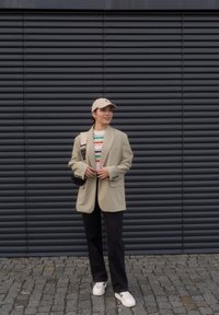 Blazer oversized em bege claro, suéter de malha estampado em várias cores, calças escuras, ténis brancos e um boné de cor clara.