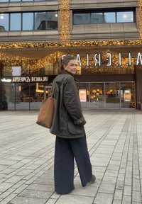 Oversized olijfgroene jas met bruine accenten, marineblauwe wijde broekspijpen en een bruine draagtas; buiten met een verlicht gebouw op de achtergrond.