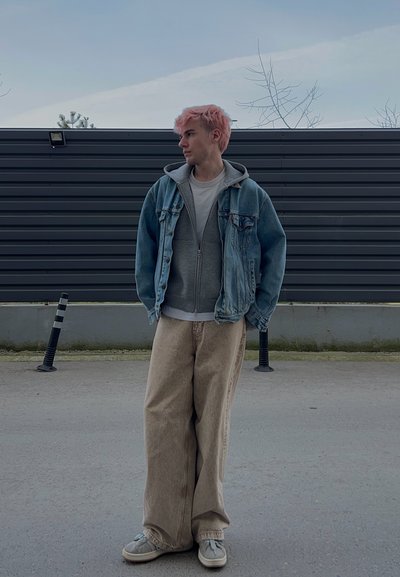 Persona joven con cabello rosa que lleva una chaqueta de mezclilla, sudadera gris con capucha, pantalones anchos beige y zapatillas, de pie sobre un suelo pavimentado cerca de una cerca oscura.