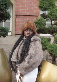 Fuskpälsjacka i ljusbrunt med huva, strukturerad finish, kombinerad med en brun pälsmössa. Modellen håller en brun handväska framför grönska.