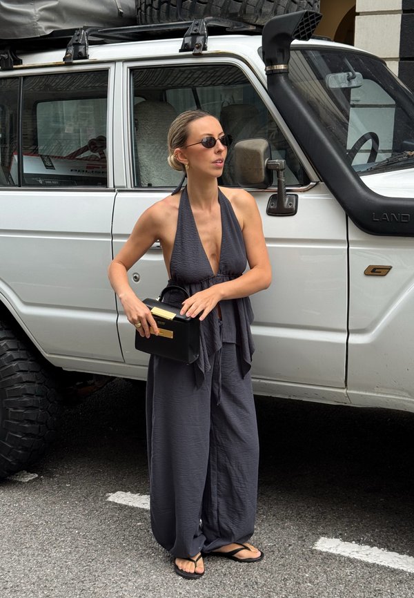 Femme en combinaison dos nu foncée et lunettes de soleil tenant un sac à main noir, debout à côté d'un véhicule tout-terrain blanc dans une rue de la ville.