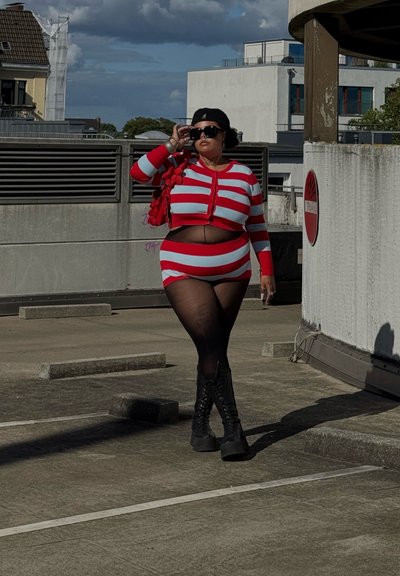 Conjunto de top corto a rayas rojas y azul claro y falda; botas negras con cordones; gafas de sol de gran tamaño; llevando un bolso texturizado rojo; de pie en un entorno urbano.