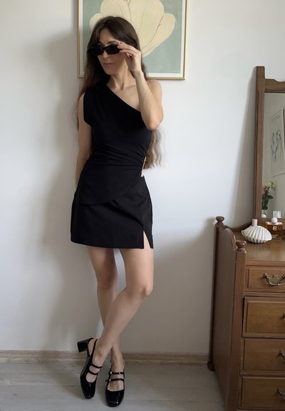 Vestido negro asimétrico con un corpiño ajustado y una abertura lateral. La modelo lleva gafas de sol negras y tacones de charol negros con tiras.