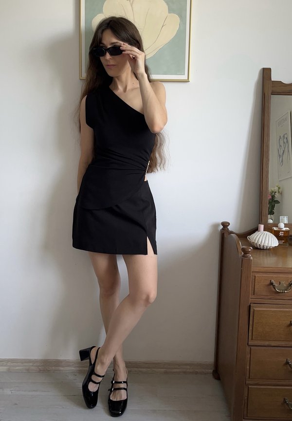 Černé jedno-ramenné šaty s přiléhavým živůtkem a bočním rozparkem. Modelka má na sobě černé sluneční brýle a lakované černé lodičky se strapci.