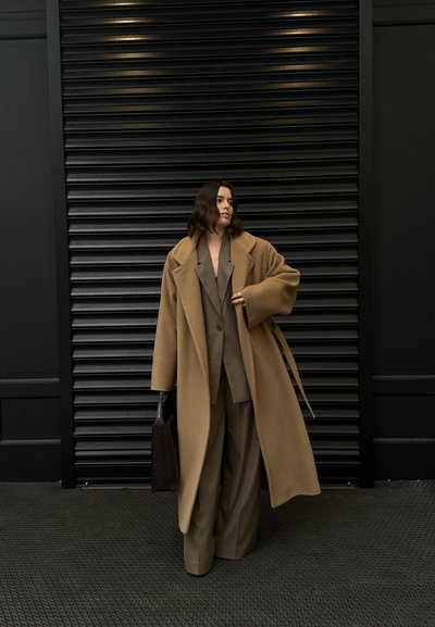 Abrigo de camello oversize sobre un traje marrón con pantalones de pierna ancha, acompañado de un bolso oscuro. El fondo presenta una superficie texturizada en negro.