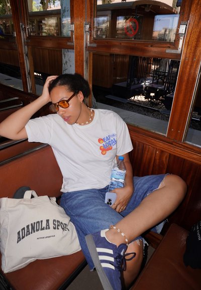 Camiseta blanca con el colorido texto "Come Local", pantalones cortos de mezclilla, gafas de sol y zapatillas azules. Sostiene una botella de agua con etiqueta visible.