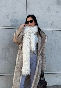 Manteau en fausse fourrure beige avec une longue écharpe blanche texturée. Haut et pantalon gris, accessoirisés de lunettes de soleil foncées et d'un sac à main marron.