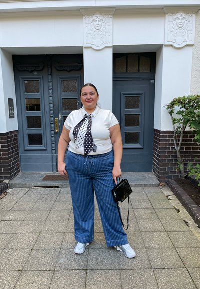 Camisa blanca de manga corta con una corbata de lunares, pantalones anchos de rayas azules y zapatillas blancas. Sosteniendo un pequeño bolso negro.