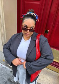 Μαύρο σακάκι με ραφή πάνω από λευκή μπλούζα, γκρι φουστάνι με ρίγες, κόκκινη τσάντα και πολύχρωφο φουλάρι σε κότσο, μπροστά από μια κόκκινη πόρτα.