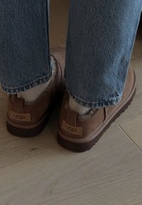 Pruunid seemisnahast libisevad jalanõud pehme sisuga, kummist tallaga ja pruun UGG logo plaastriga kandadel, kombineeritud teksapükstega.