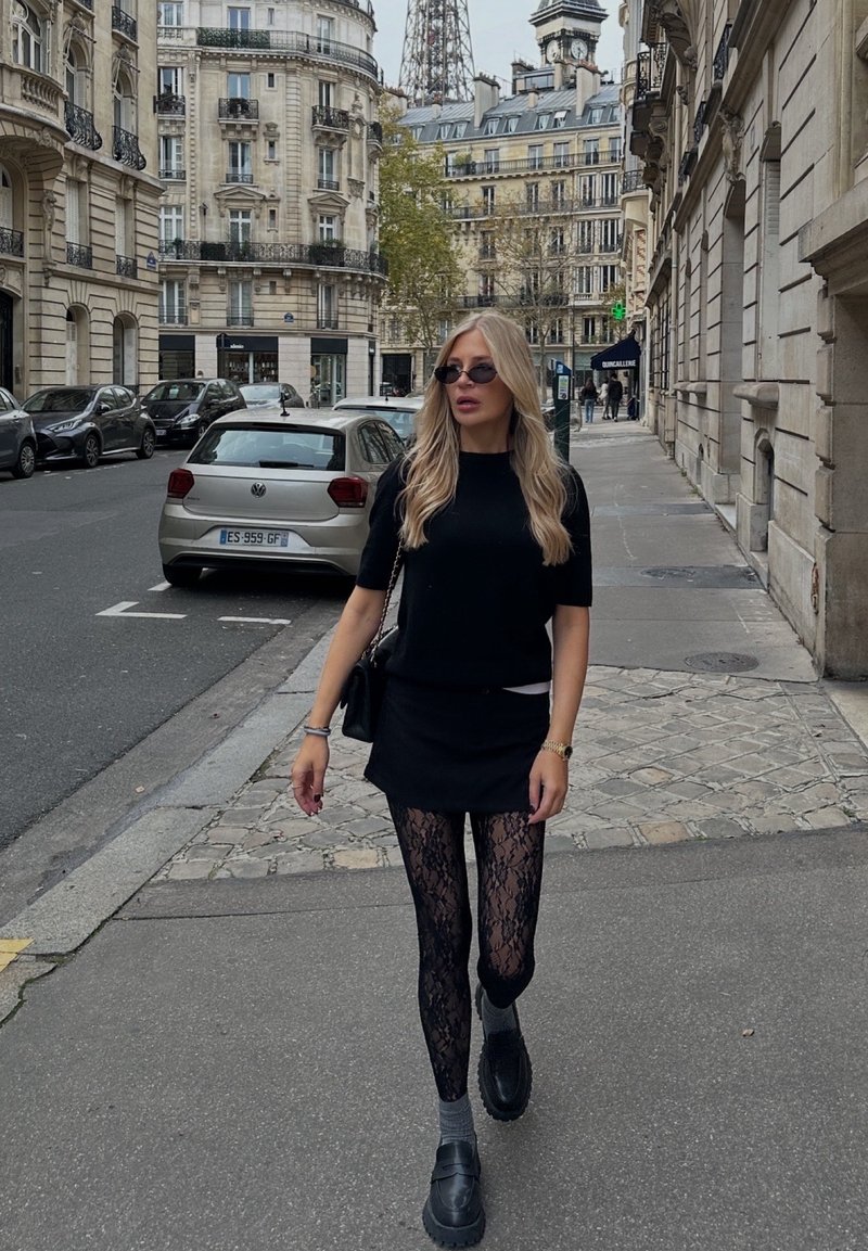 Femme en haut en maille noire, mini-jupe et collants en dentelle marchant dans une rue parisienne. Elle porte des lunettes de soleil et un sac à main noir.