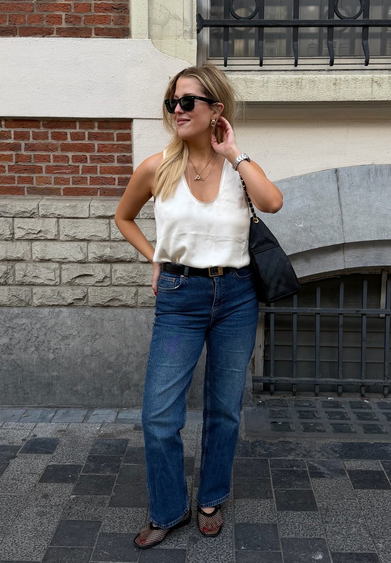 Blond kvinne med solbriller, hvit ermeløs topp, blå jeans, svart belte, nett sko og bærer en svart skulderveske på en fortau i byen.