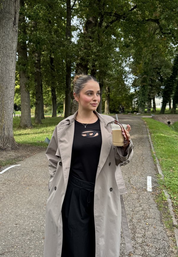 Beige trench coat over black top with cut-out detail, black pants, holding a glass of light brown drink with a straw, in a park setting.