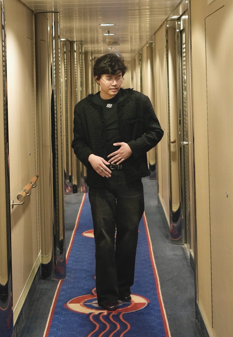 Jeune personne avec des lunettes en vêtement noir marchant dans un couloir étroit avec un tapis bleu et rouge à motifs.