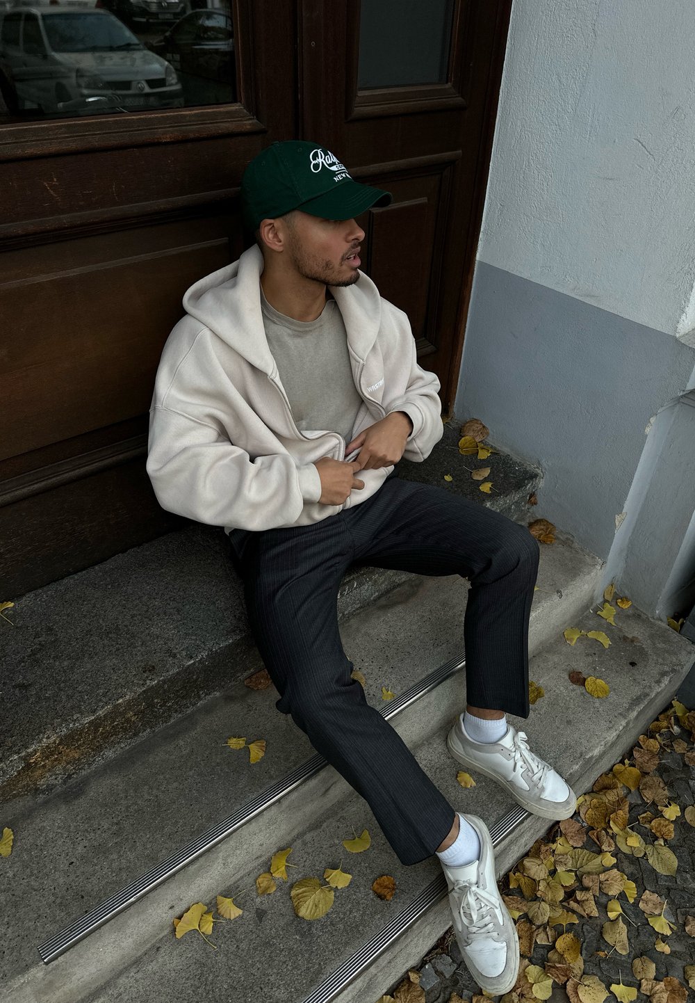 Κρεμ φούτερ με φερμουάρ, γκρι μπλούζα από κάτω, σκούρα παντελόνια με ρίγες και λευκά αθλητικά παπούτσια. Καθισμένος σε σκαλιά με φθινοπωρινά φύλλα γύρω.