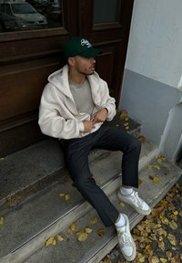 Κρεμ φούτερ με φερμουάρ, γκρι μπλούζα από κάτω, σκούρα παντελόνια με ρίγες και λευκά αθλητικά παπούτσια. Καθισμένος σε σκαλιά με φθινοπωρινά φύλλα γύρω.