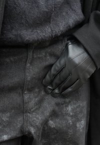 Gant en cuir noir reposant sur la taille d'un jean en denim délavé acide foncé, associé à un pull foncé en matière pelucheuse et à une manche de manteau noire.