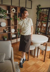 Crème polo shirt met zwarte afwerking; bruine shorts; witte sokken; zwarte schoenen. Binnenomgeving met houten meubels en planten. Ronde tafel.
