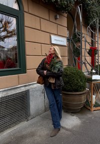Veste en cuir marron, jean bleu, bottines grises ; tenant des branches de pin vertes avec des baies rouges. Devanture de magasin avec décorations de fenêtres.