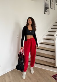 Femme portant un soutien-gorge de sport noir, une veste noire, un pantalon de survêtement rouge, des baskets blanches, tenant un sac de sport noir, debout près d'un escalier en bois à l'intérieur.