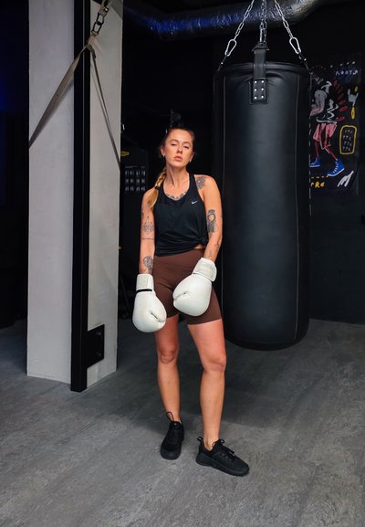 Guantes de boxeo negros y una bolsa de entrenamiento negra en un gimnasio. La persona lleva una camiseta sin mangas negra y pantalones cortos marrones, con zapatillas negras.