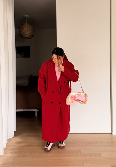 Persona con un abrigo largo rojo, camisa rosa y zapatillas, sosteniendo un bolso rosa y naranja, de pie en una habitación minimalista con suelo de madera.