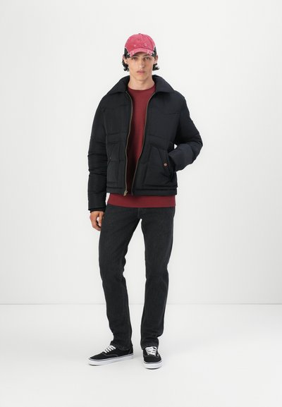 Chaqueta de plumas negra con dos bolsillos frontales y cierre de cremallera, llevada sobre un suéter burdeos y jeans negros, complementada con una gorra rosa.