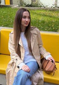 Jovem mulher com um trench coat bege e um suéter branco sentada num banco amarelo ao ar livre, segurando uma bolsa castanha, olhando para a direita.