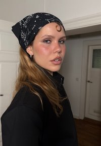 Czarna bandana z białymi wzorami, noszona w związanym stylu. Model w czarnej bluzie na zamek, z długimi, falowanymi włosami częściowo widocznymi.
