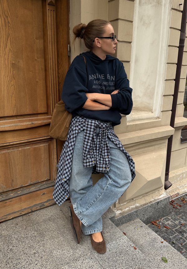 Mujer con moño, con sudadera negra, vaqueros claros y camisa de cuadros atada a la cintura, está de pie junto a un edificio cerca de una puerta de madera.