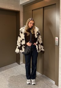 Mehko črno-belo krzno, rjav pulover, temne kavbojke in sivi supergi. Stoji pred dvigalom v sodobnem okolju.